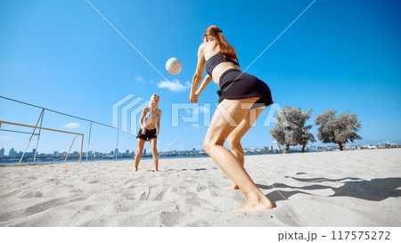 Two girls, friends in beach wear playing beach volleyball under clear blue sky and urban building on background. Heated beach match Two girls, friends in beach wear playing beach volleyball under clear blue sky and urban building on background. Heated beach match 117575272