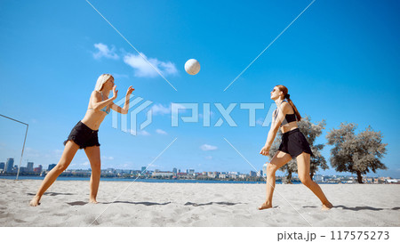 Two girls, friends in beach wear playing beach volleyball under clear blue sky and urban building on background. Heated beach match 117575273