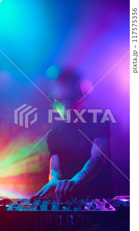 DJ immersed in vibrant lights, mixing beats as colors pulse around him, creating electrifying atmosphere. Dancefloor. 117575356