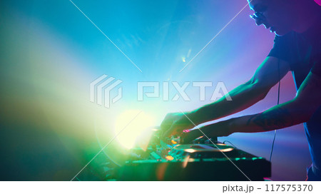 DJ stands in vibrant spotlight, surrounded by vibrant light show, his music shaping electric energy of scene. 117575370