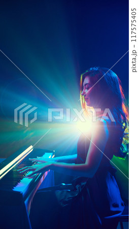 Pianist's music seems to paint air with colors of light that swirl around her, fusion of sound and vision. Woman playing piano on stage. Pianist's music seems to paint air with colors of light that swirl around her, fusion of sound and vision. Woman playing piano on stage. 117575405
