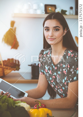 An attractive young dark-haired woman chooses the recipe for a delicious meal while sitting at the table in sunny kitchen An attractive young dark-haired woman chooses the recipe for a delicious meal while sitting at the table in sunny kitchen 117575572