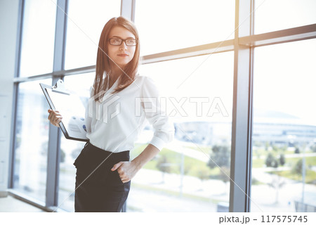 Beautiful female specialist with clipboard standing in sunny office and smiling charmingly. Business people concept 117575745