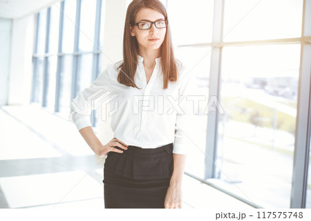 Beautiful female specialist with dark hair standing in sunny office and smiling charmingly. Working on design, data analysis, plan strategy Beautiful female specialist with dark hair standing in sunny office and smiling charmingly. Working on design, data analysis, plan strategy 117575748