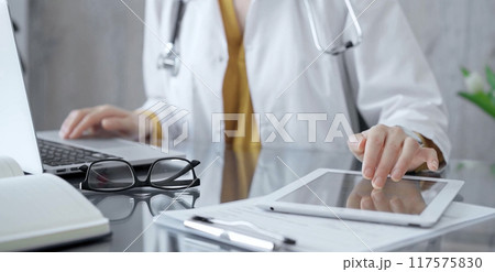 Doctor woman using tablet on the glass desk in medical office. Close-up of a doctor's hands interacting with a touch pad in clinic. Medicine and health care 117575830