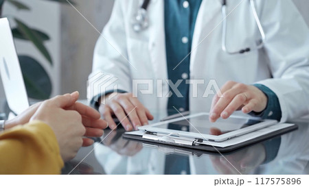 Doctor and patient consultation in clinic. Patient woman is gesturing over a glass desk while a physician is using tablet computer making some notes, close up of hands. Medicine and health care 117575896