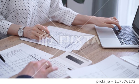 Female bookkeeper is working on finances papers with a colleague, using magnificent glass, calculator and laptop. Audit, taxes 117576172