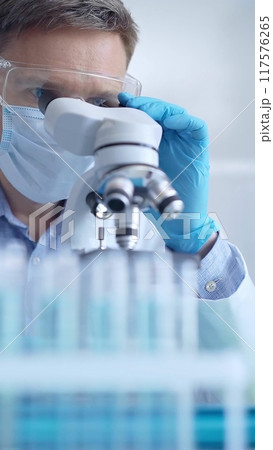 Man scientist with protective glasses, mask and gloves is using microscope in laboratory, vertical portrait of researcher. Microbiology and science Man scientist with protective glasses, mask and gloves is using microscope in laboratory, vertical portrait of researcher. Microbiology and science 117576265