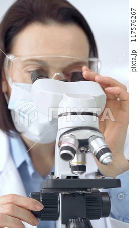 Female scientist using microscope in laboratory. Focus on a woman in lab goggles analyzing samples with a microscope in a scientific setting 117576267