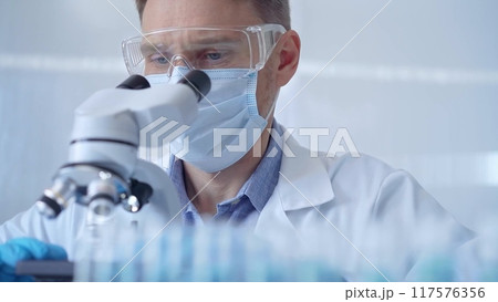 Man scientist with protective glasses, mask and gloves is using microscope in laboratory, portrait of researcher. Microbiology science concept Man scientist with protective glasses, mask and gloves is using microscope in laboratory, portrait of researcher. Microbiology science concept 117576356
