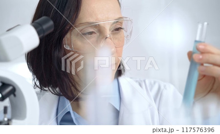 Female scientist analyzing sample in laboratory. Focused female researcher examines a clear test tube in a modern lab setting Female scientist analyzing sample in laboratory. Focused female researcher examines a clear test tube in a modern lab setting 117576396