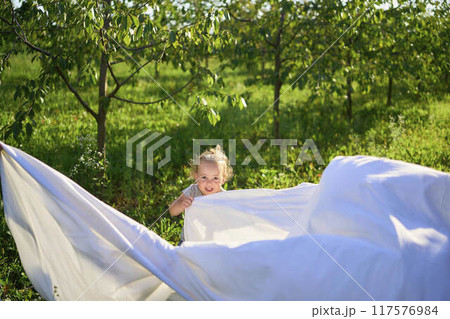 a family of four is playing with a white sheet in the garden, a toddler wants to flutter the sheet 117576984
