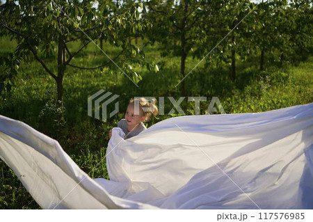 a family of four is playing with a white sheet in the garden, a toddler wants to flutter the sheet 117576985