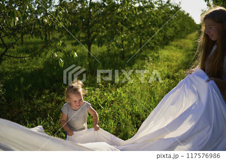 a family of four is playing with a white sheet in the garden, a toddler wants to flutter the sheet 117576986