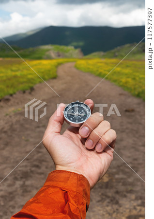 Compass in hand against the background of a mountain road, vertical photo. Choosing a path in travel and hiking 117577397