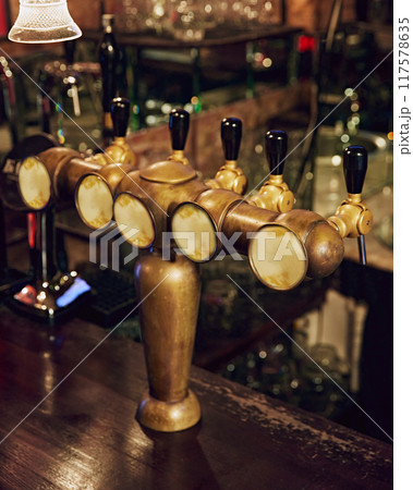 Close-up of row of shiny beer taps with handles and mirrors, set against dark background with blurred bar counter and empty glasses in background. Close-up of row of shiny beer taps with handles and mirrors, set against dark background with blurred bar counter and empty glasses in background. 117578635