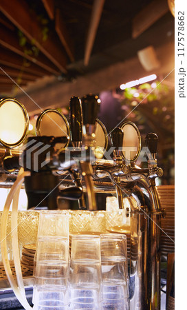 Row of golden shiny beer taps with handles, set against dark background with empty glasses, with a wooden ceiling and warm lighting in the background. Row of golden shiny beer taps with handles, set against dark background with empty glasses, with a wooden ceiling and warm lighting in the background. 117578662