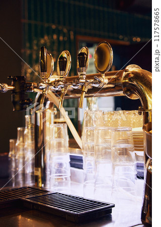 Close-up of shiny beer tap with multiple handles, set against dark background with empty glasses and metal grid in the foreground. 117578665