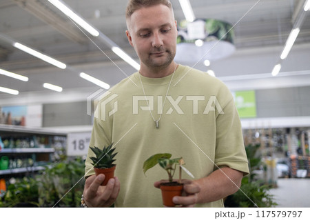 Portrait of smiling young bearded man in green t shirt holding while choosing plants in home goods store. Plant, nature, shopping concept Portrait of smiling young bearded man in green t shirt holding while choosing plants in home goods store. Plant, nature, shopping concept 117579797