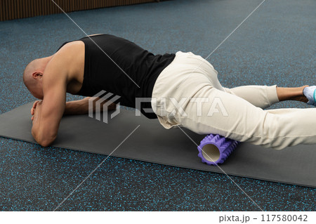 Athlete man exercising and stretching legs on foam roller on sports ground. Outdoor activity. Morning routine. Public park. Athlete man exercising and stretching legs on foam roller on sports ground. Outdoor activity. Morning routine. Public park. 117580042