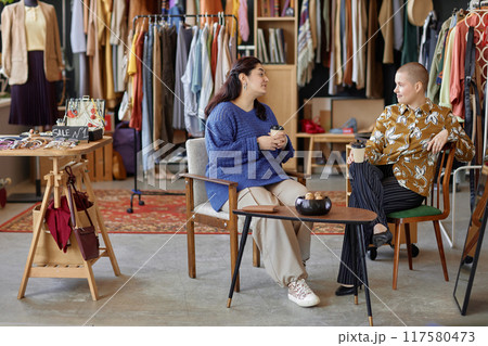 Wide angle shot of two Gen Z female friends sitting at tea table in vintage chairs chatting over cup of coffee discussing fashion at thrift store, copy space Wide angle shot of two Gen Z female friends sitting at tea table in vintage chairs chatting over cup of coffee discussing fashion at thrift store, copy space 117580473