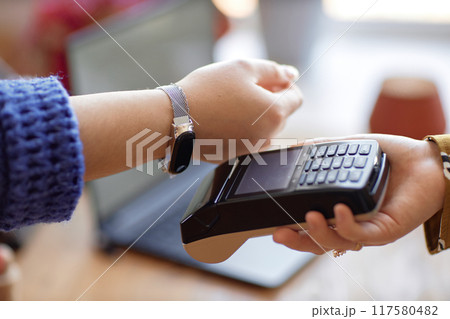 Close up on on female hand paying wireless with smartwatch and unrecognizable cashier using POS terminal at store, copy space Close up on on female hand paying wireless with smartwatch and unrecognizable cashier using POS terminal at store, copy space 117580482