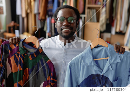 Portrait shot of smiling Black man in vintage glasses holding wooden hangers with second hand shirts while looking at camera at thrift store 117580504