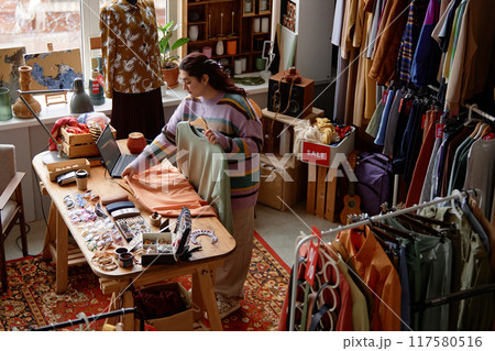 Wide angle shot of female sales assistant working at checkout desk cluttered with fashion accessories at second hand shop with retro style carpet selling clothes and household goods, copy space 117580516