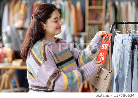 Side view medium shot of smiling Middle Eastern female customer choosing to buy second hand purse with fifty percent discount tag at thrift store Side view medium shot of smiling Middle Eastern female customer choosing to buy second hand purse with fifty percent discount tag at thrift store 117580634