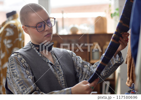 Medium shot of young fashionable female shopper with buzz cut choosing garment picking up from second hand clothes at thrift store, copy space Medium shot of young fashionable female shopper with buzz cut choosing garment picking up from second hand clothes at thrift store, copy space 117580696