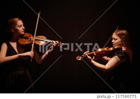 Side view of elegant string duet of female violinists playing classical music in contrast top light on stage with black curtains, copy space 117580773