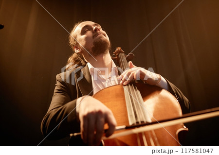 Low angle view of male musician with eyes closed passionately playing cello against black curtains in studio with warm contrast light, copy space 117581047