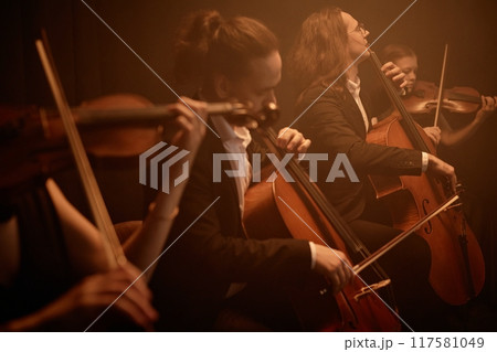 String quartet passionately playing cellos and violins while sitting in row on stage in misty orange light, copy space 117581049