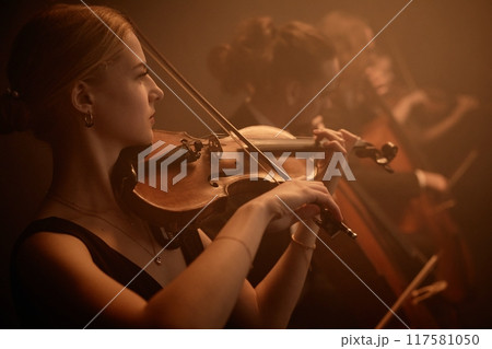 Side portrait of virtuoso female classic music artist playing violin with string quartet on stage in misty orange light 117581050