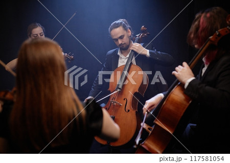Young male musician in black suit playing cello with professional string quartet in hazy dark studio with blue top light 117581054