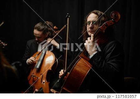 Concentrated male cello player in glasses performing classical music with string quartet in studio with dramatic top light against black background Concentrated male cello player in glasses performing classical music with string quartet in studio with dramatic top light against black background 117581057