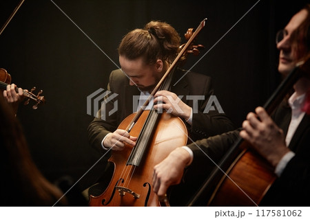 Medium shot of male musician looking down, while strumming cello playing classical music with dedication in string ensemble in studio with contrast top light on black background 117581062