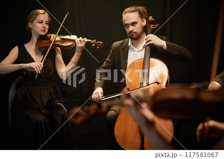 Virtuoso female and male string musicians playing violin and cello on stage with contrast top light against black curtains Virtuoso female and male string musicians playing violin and cello on stage with contrast top light against black curtains 117581067