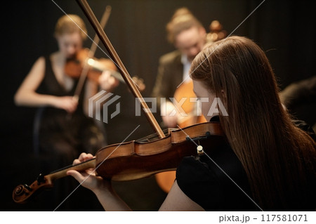Rear view of unrecognizable female orchestra musician playing violin with string quartet in spotlight on stage with black curtains Rear view of unrecognizable female orchestra musician playing violin with string quartet in spotlight on stage with black curtains 117581071