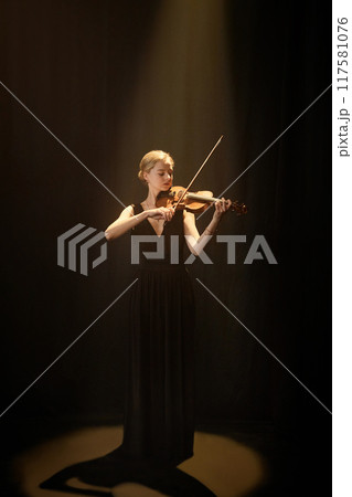 Vertical full shot of concentrated female violin player performing solo concerto under dramatic projector light in dark studio on black background, copy space 117581076