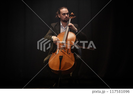 Full shot of male classical music performer playing cello during solo concert while sitting in chair against black curtains under projector light, copy space 117581096