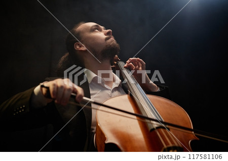 Low angle view of moved male musician thoughtfully looking up while playing cello in hazy dark studio with top light, copy space 117581106