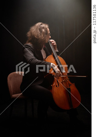 Vertical full shot of male cello player in black tuxedo performing classical music while sitting in chair in dark studio with top light, copy space Vertical full shot of male cello player in black tuxedo performing classical music while sitting in chair in dark studio with top light, copy space 117581136