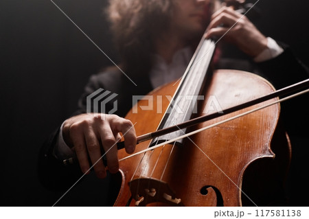 Cropped shot of unrecognizable talented male cello player performing classical music in dark recording studio with dim light, focus on violoncello bow Cropped shot of unrecognizable talented male cello player performing classical music in dark recording studio with dim light, focus on violoncello bow 117581138