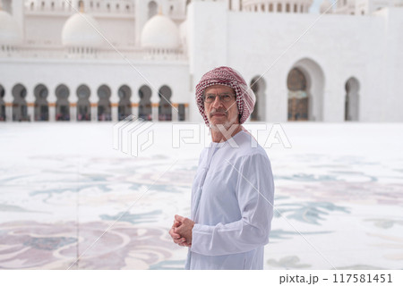 Man in traditional attire poses outside grand mosque with domes and arches. 117581451
