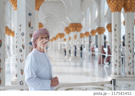 Man in traditional attire stands inside ornate mosque with columns behind. Man in traditional attire stands inside ornate mosque with columns behind. 117581452