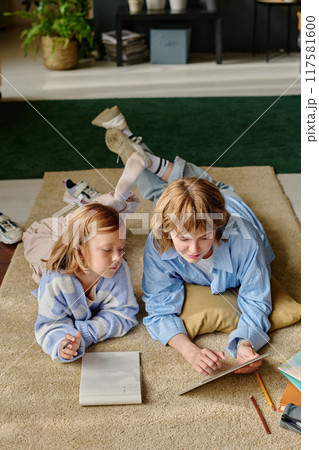 High angle shot of young Caucasian mom holding tablet and showing presentation to her daughter while they lying on carpet 117581600