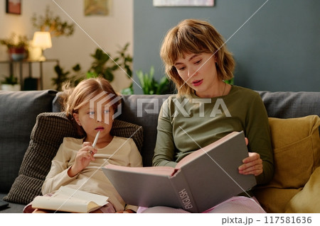 Lovely mom holding huge book with gray cover and reading aloud while her little girl listening to fairytale Lovely mom holding huge book with gray cover and reading aloud while her little girl listening to fairytale 117581636