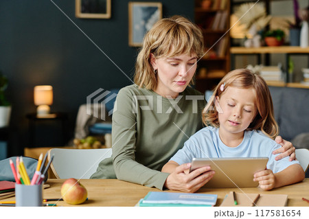 Mom and her daughter with similar haircuts sitting at wooden desk and watching cartoons via tablet together Mom and her daughter with similar haircuts sitting at wooden desk and watching cartoons via tablet together 117581654