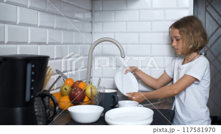 Young girl washing dishes in the kitchen sink, dressed in a white t-shirt, household duties 117581970